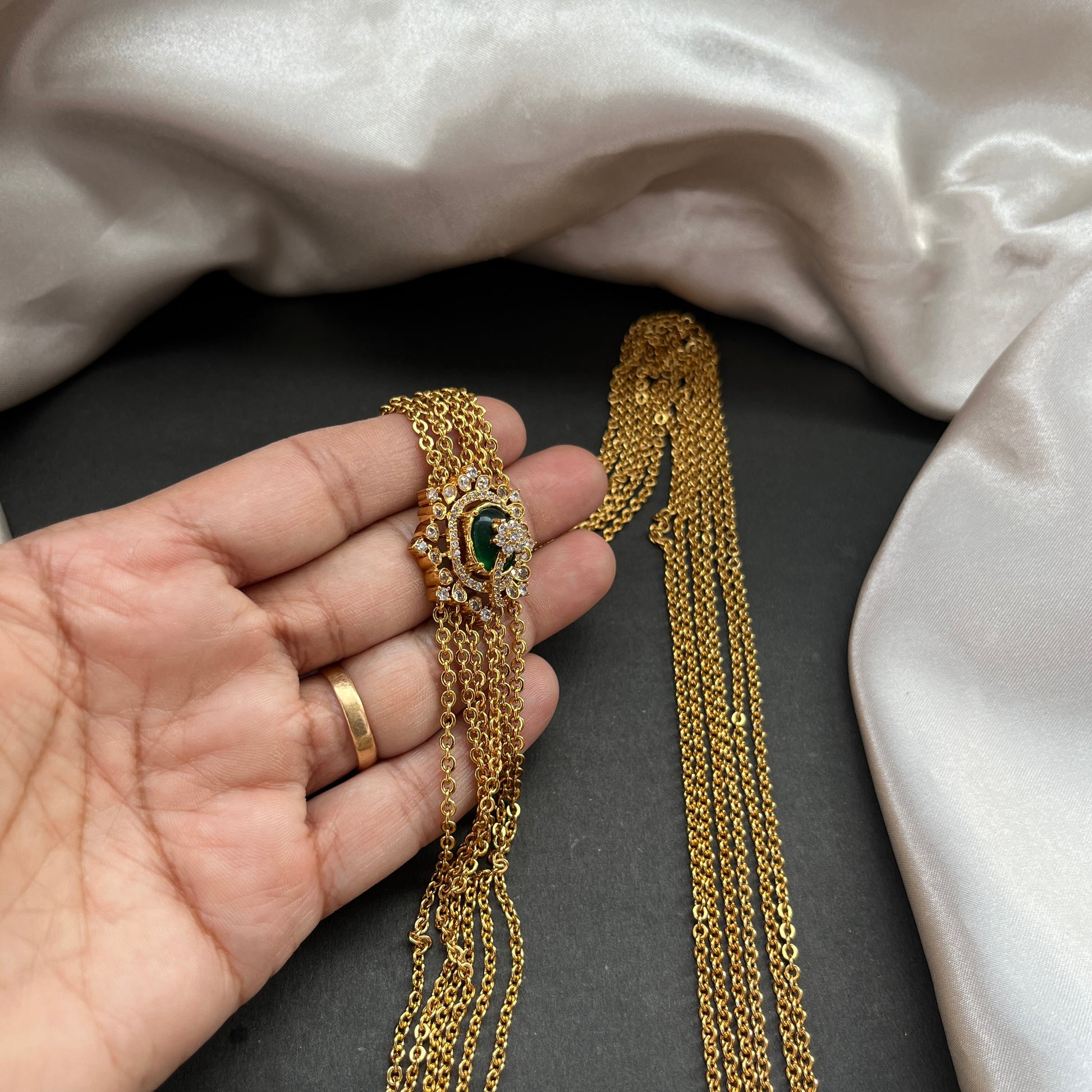 Gold necklace with green gemstone held by a hand on a dark surface with a light fabric background